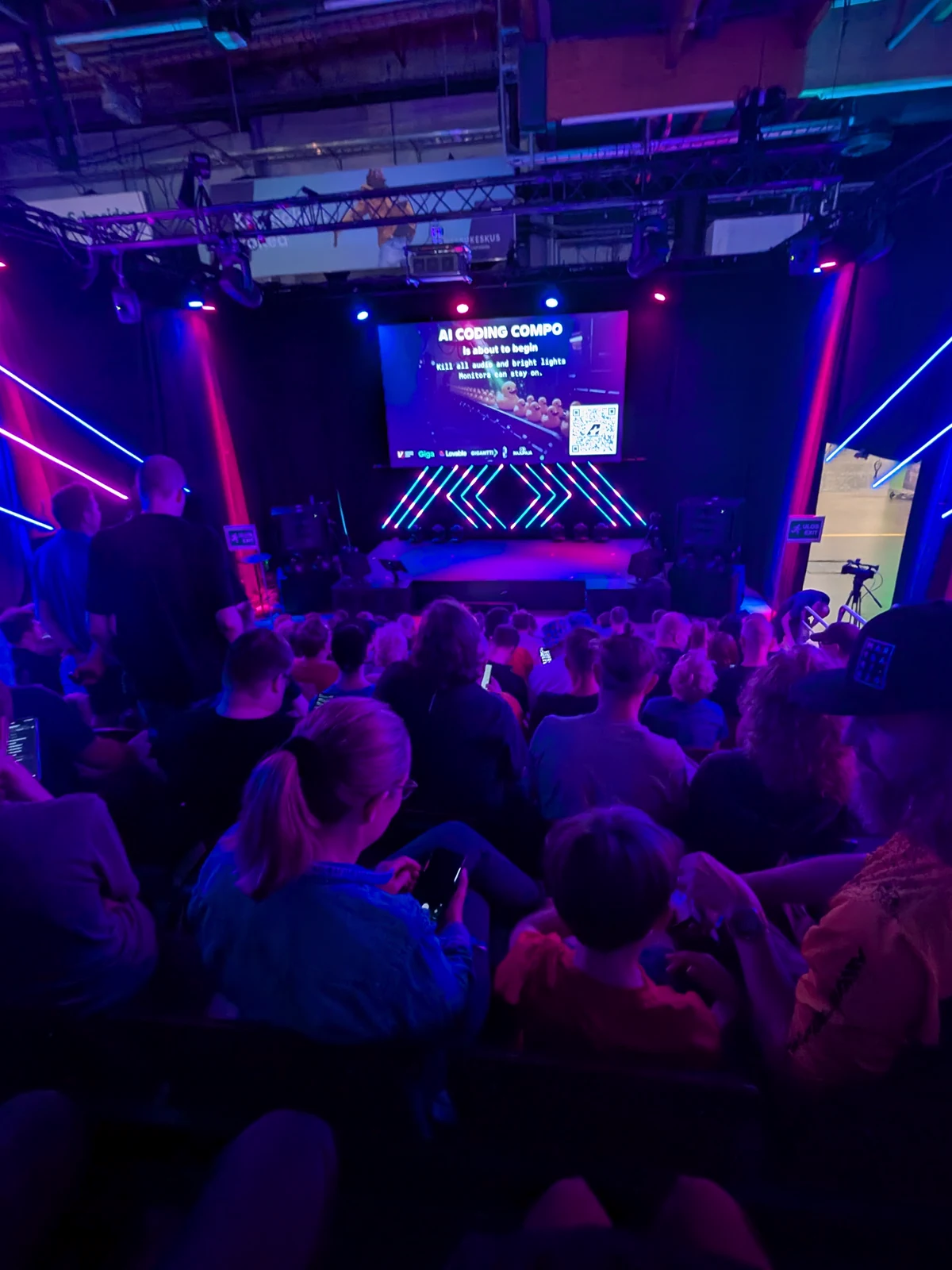 A full venue under blue and magenta light, the audience facing a stage screen reading AI Coding Competition